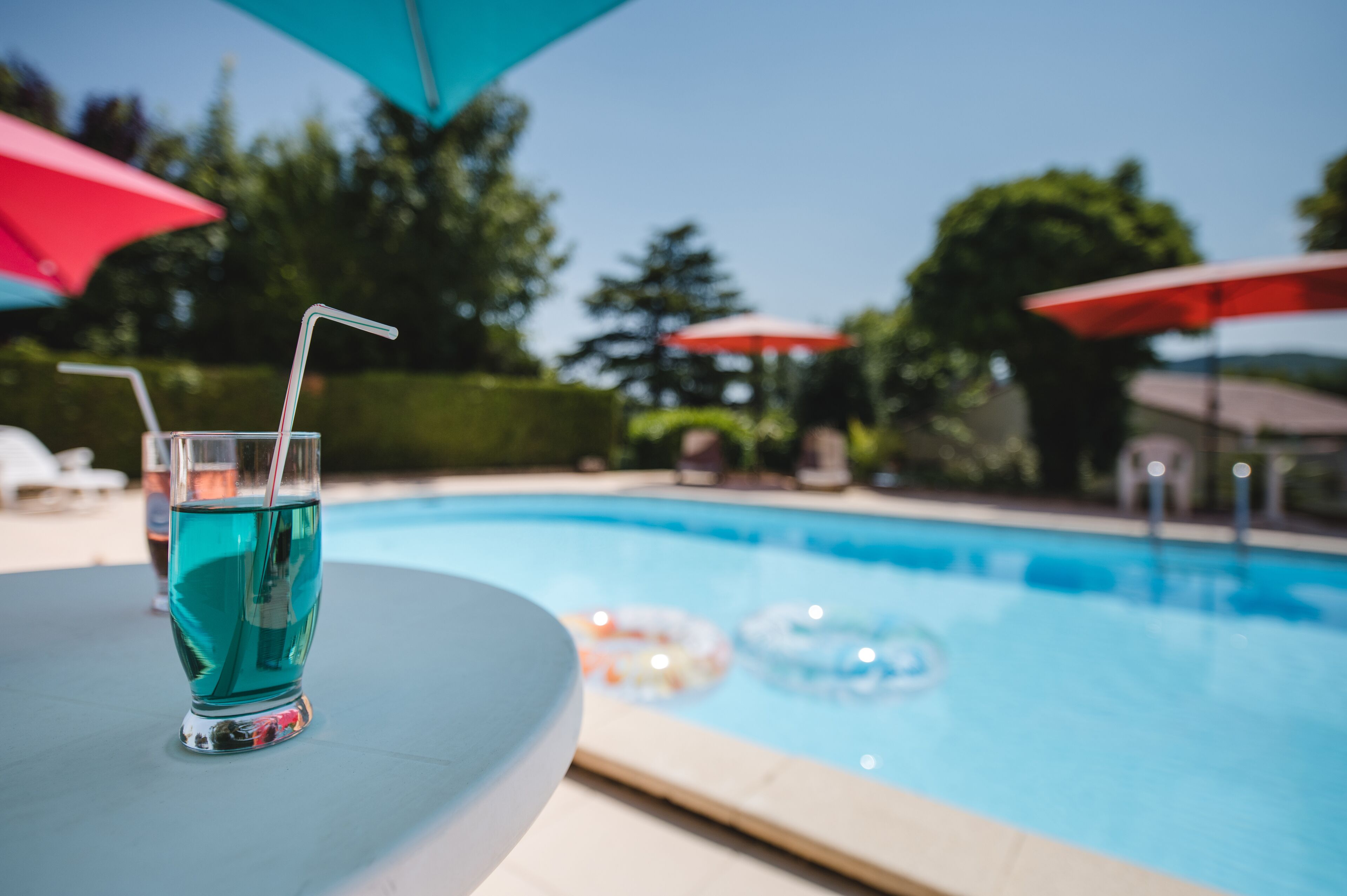 Vista clara de una piscina con un vaso de líquido azul y una pajita, rodeado de sombrillas y sillas de descanso.