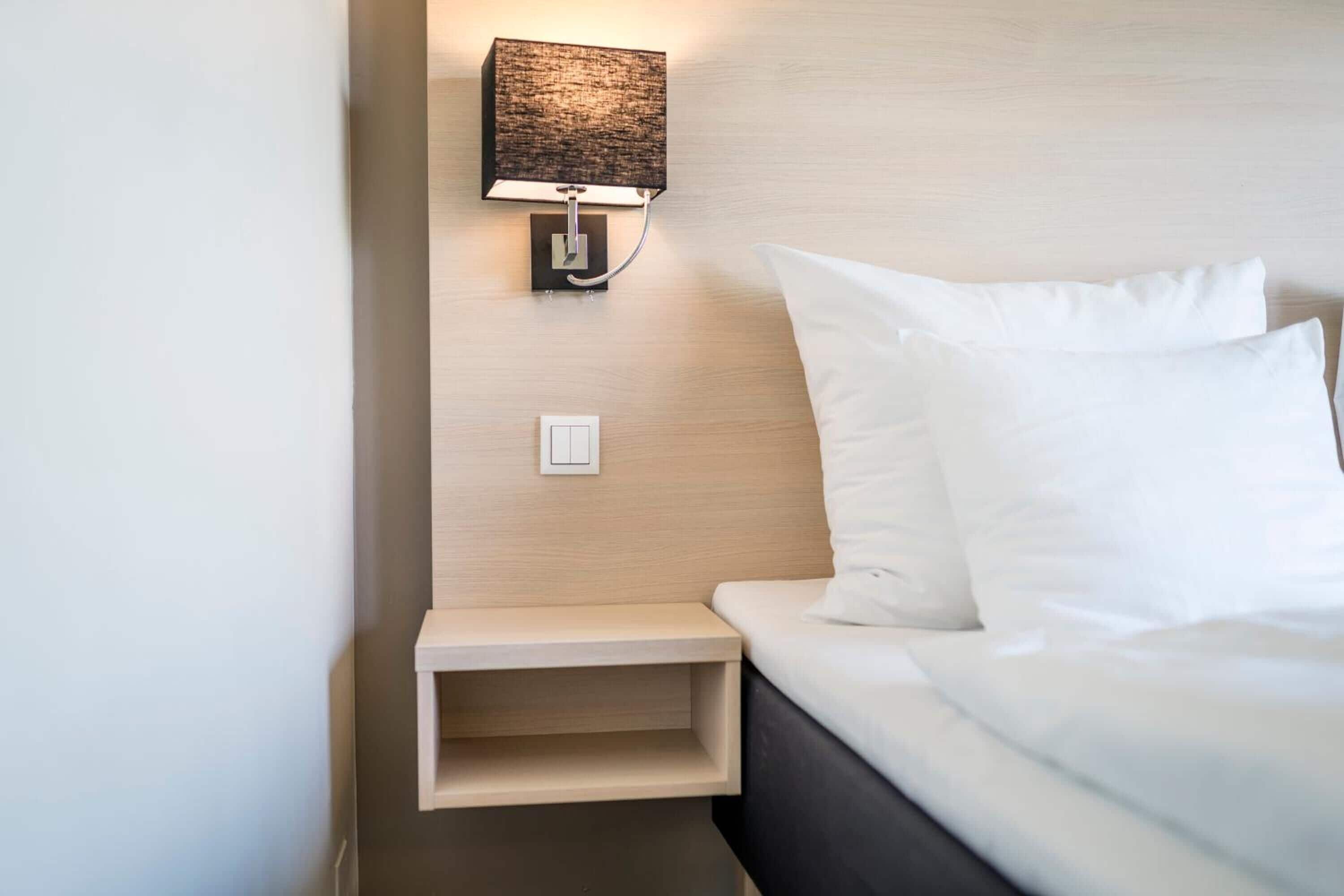 En pænt redt seng med hvide sengetøj, et natbord med lampe, og en vægmonteret lysarmatur.