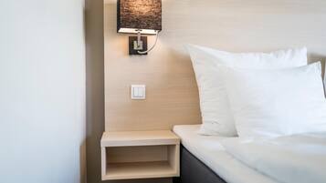 Cama bem arrumada com lençóis brancos, mesa de cabeceira com abajur e luminária na parede.