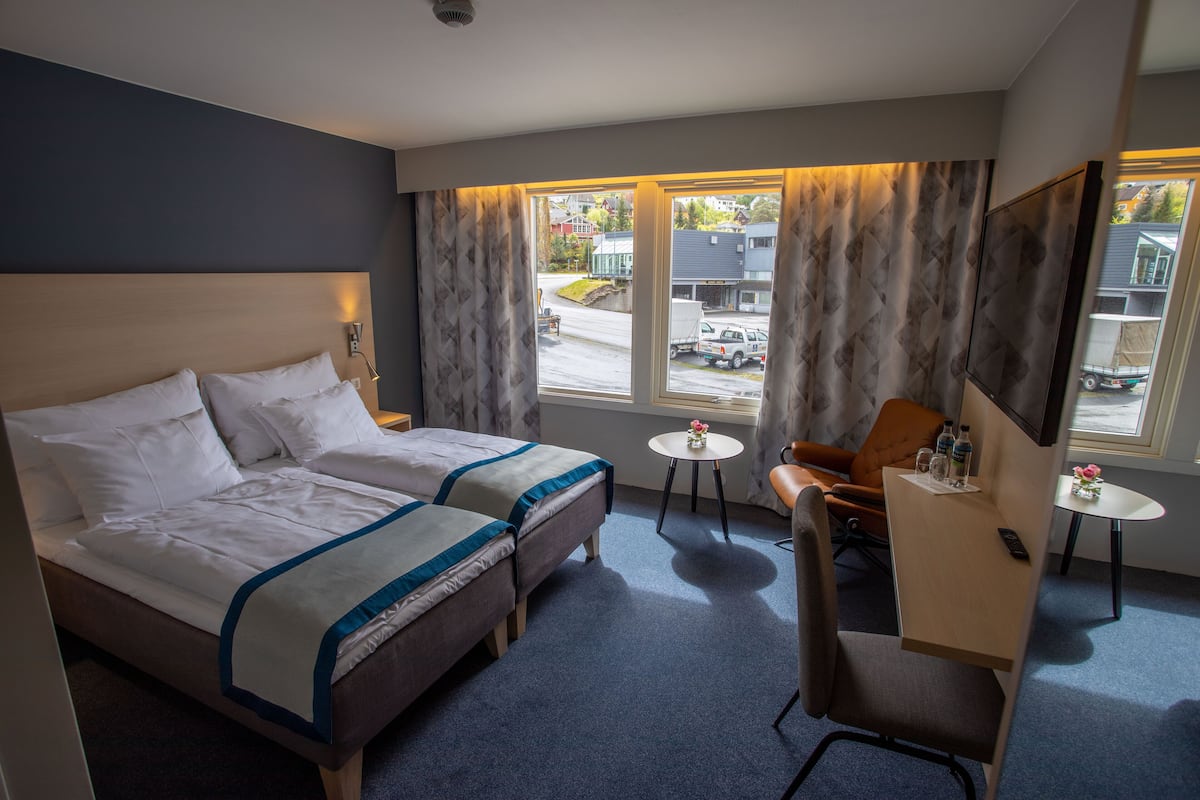 A hotel room with two beds, a desk, a chair, a small table, and a large window with curtains.