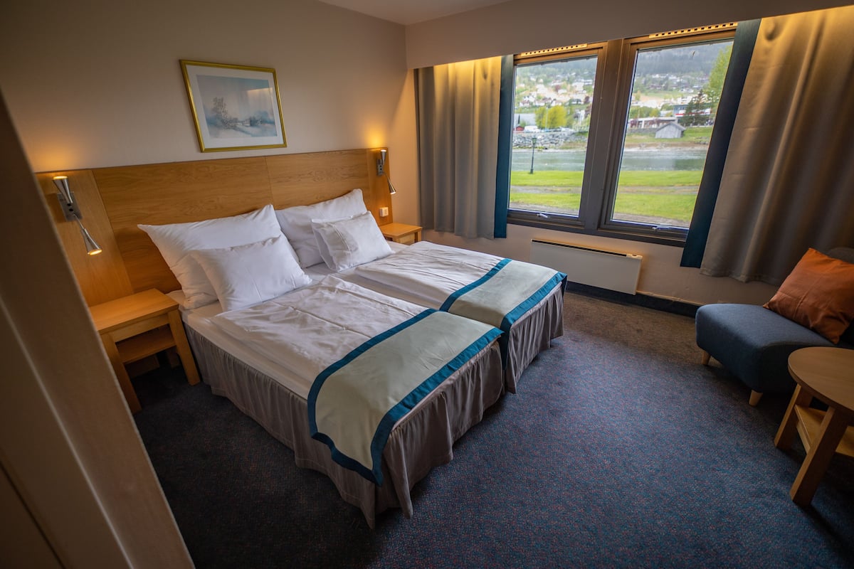 A hotel room with a large bed, a nightstand, a desk, a chair, and a window with a view of a town.