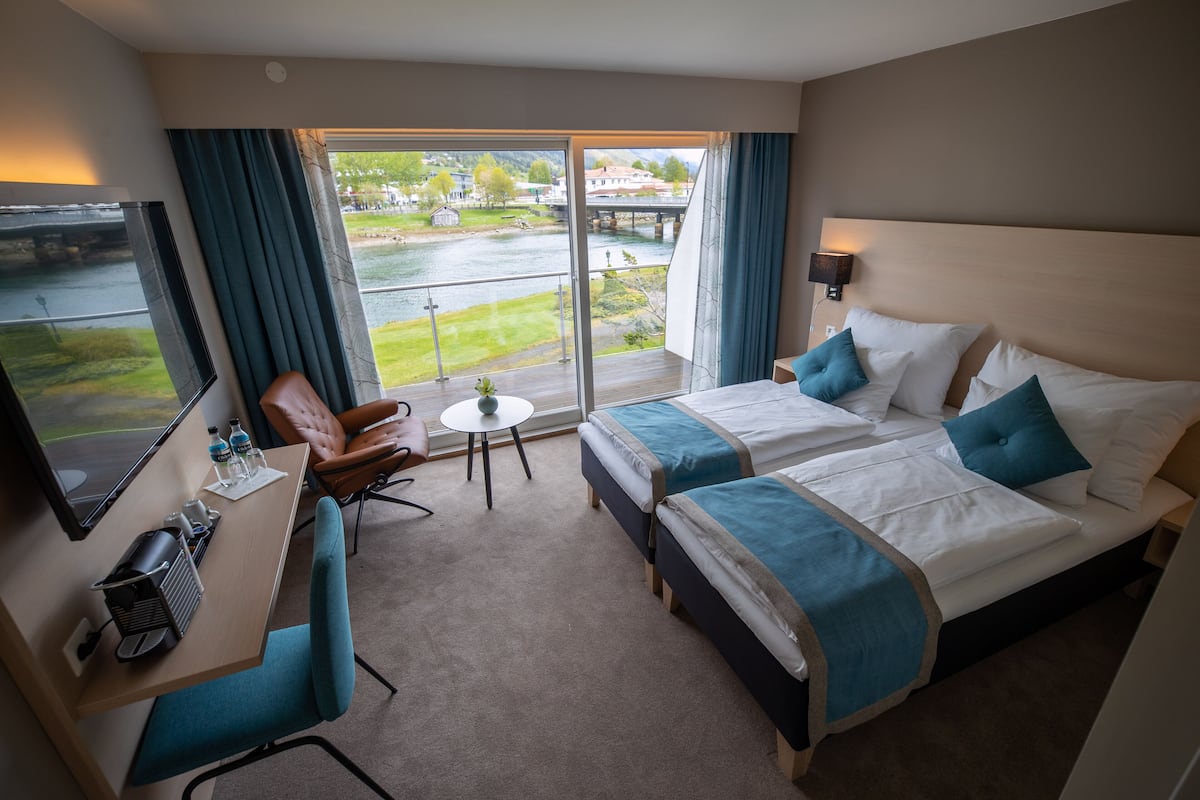 A hotel room with two beds, a desk, a chair, a small table, and a view of a river and bridge.