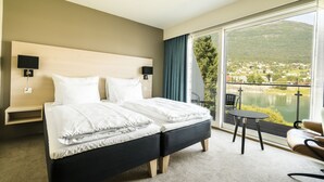 Habitación de hotel con una cama grande, dos mesitas de noche, una silla y vistas a un lago y montañas.