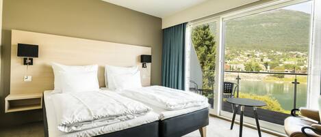 A hotel room with a large bed, two bedside tables, a chair, and a view of a lake and mountains.