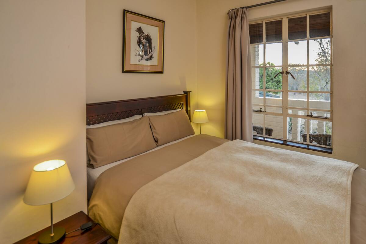 A neatly made bed with a headboard, two bedside lamps, a framed picture on the wall, and a window with curtains.