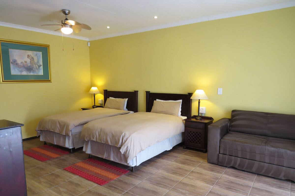 A hotel room with two beds, a sofa, a ceiling fan, and a painting on the wall.