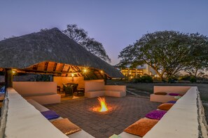 Un espacio al aire libre con techo de paja, que incluye una chimenea rodeada de asientos acolchados y un suelo empedrado.