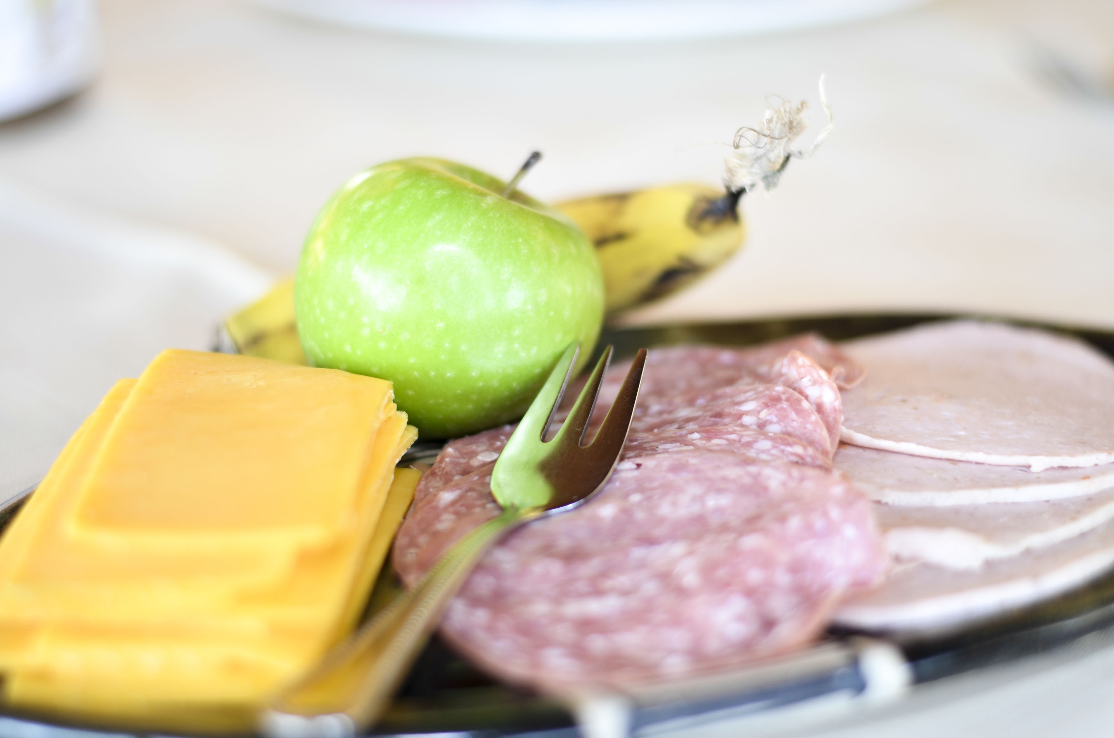 A plate with a slice of ham, a piece of cheese, a green apple, and a banana.