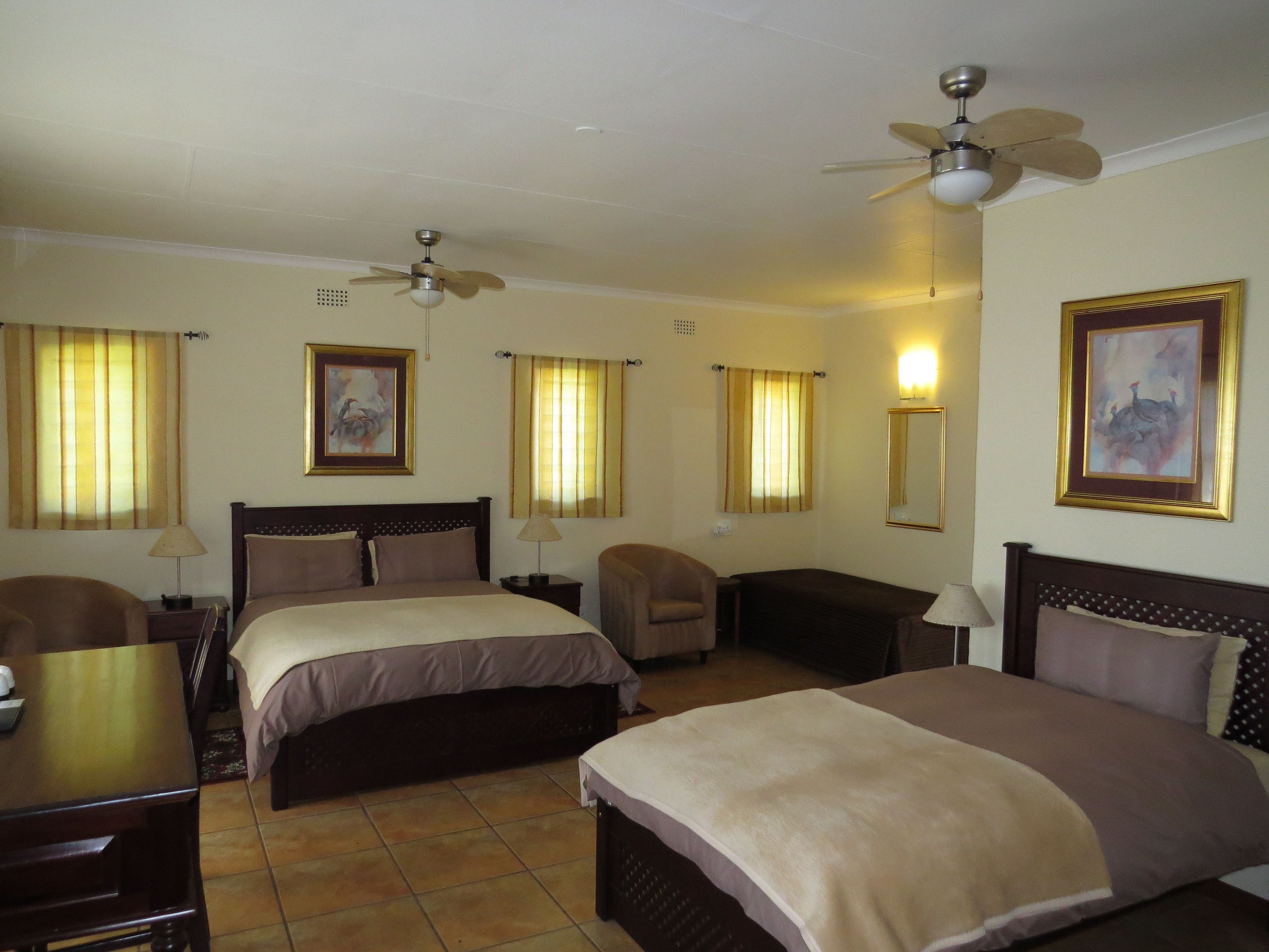 A hotel room with two beds, a desk, a chair, a ceiling fan, and a painting on the wall.