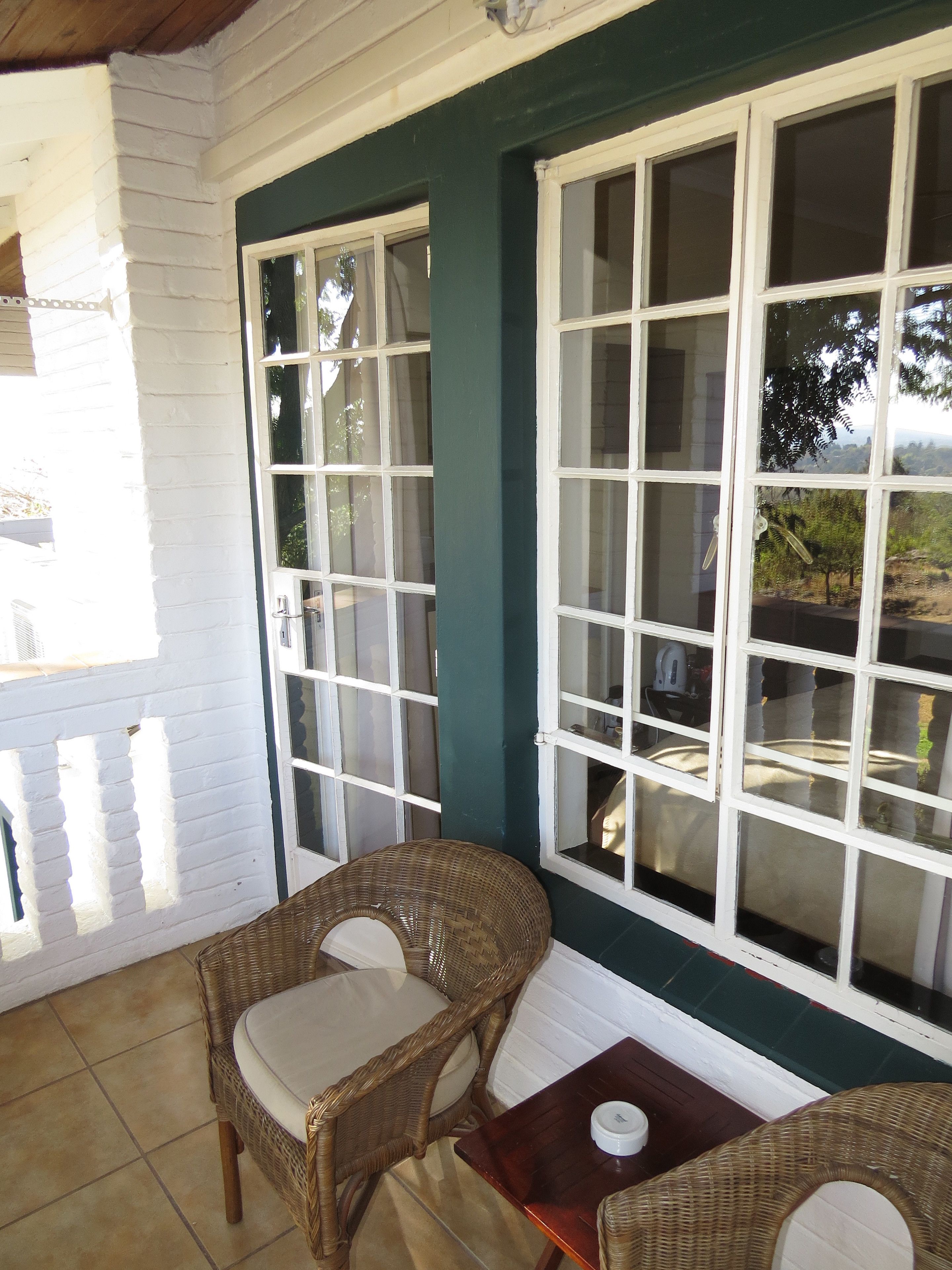 A room with two wicker chairs and a small table, large windows with white frames, and a view of greenery outside.