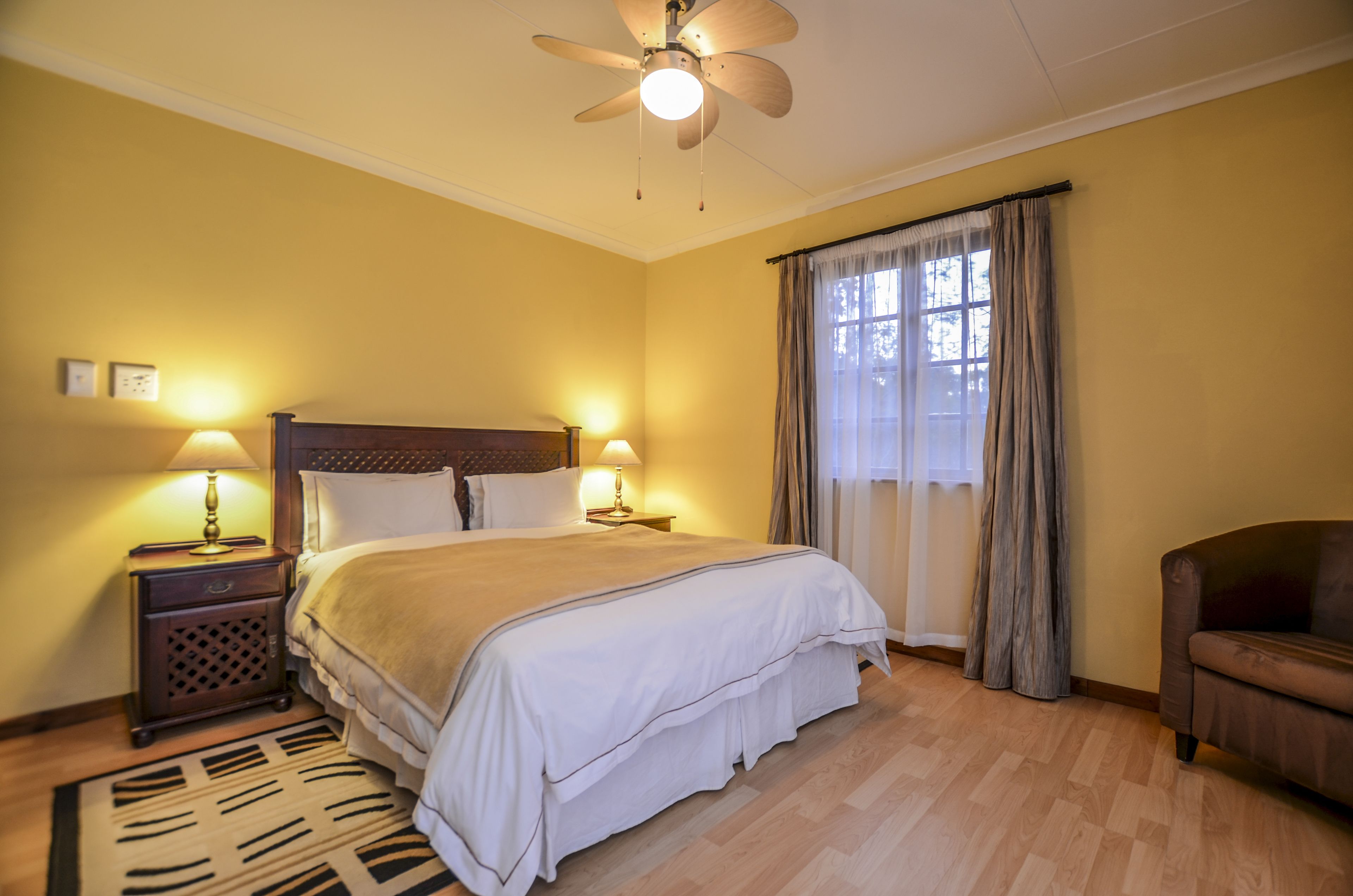 A bedroom with a bed, two bedside tables with lamps, a ceiling fan, a window with curtains, and a wooden floor.