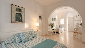 Un dormitorio con cama, mesita de noche, lámpara, un cuadro, escritorio, silla y un arco que da a otra habitación.