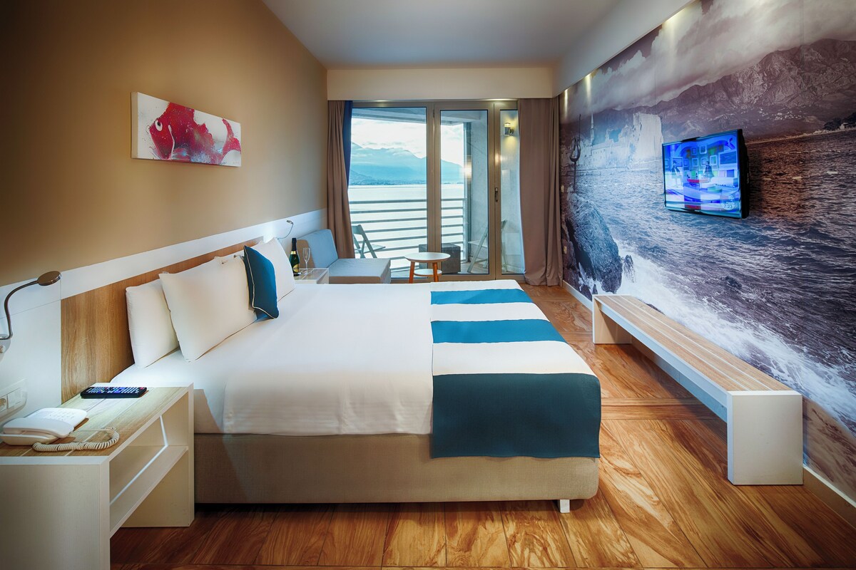 A hotel room with a large bed, a desk with a telephone, a television mounted on the wall, and a view of the sea through the window.