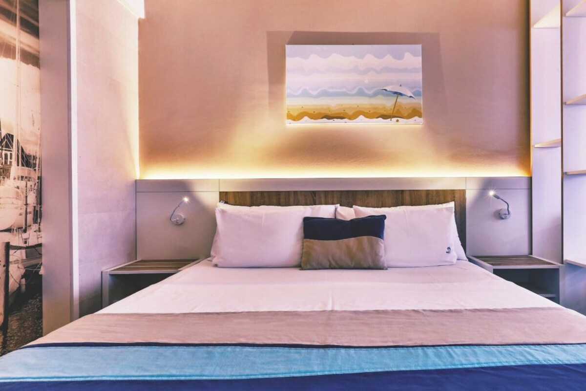 A modern hotel room with a large bed, a painting of a beach scene, and built-in shelving on the right wall.