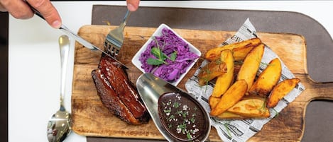 Une planche en bois sur laquelle sont disposés une assiette de travers de porc grillés, un saladier de chou rouge râpé et des quartiers de pommes de terre rôtis.