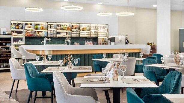 Un restaurante moderno con sillas color turquesa y blancas, una zona de bar con diversas bebidas, y una decoración interior limpia y contemporánea.
