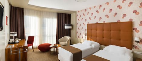 Habitación de hotel con dos camas, un escritorio, una silla, una mesita con una bandeja de frutas y un otoman rojo.