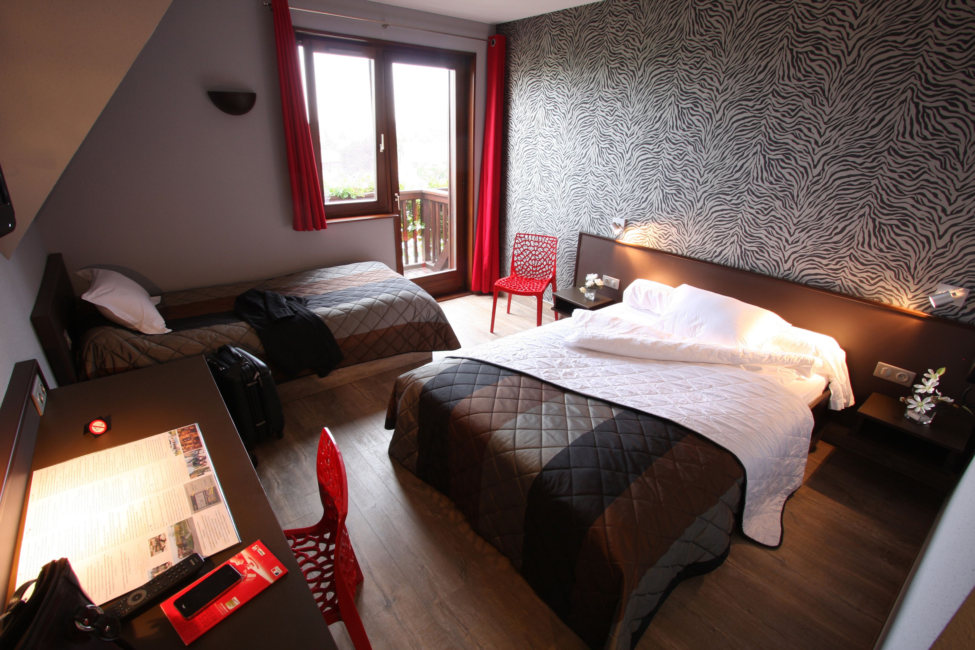 Habitación de hotel con dos camas, un escritorio, una silla, una mesita y un ventanal con cortinas rojas.