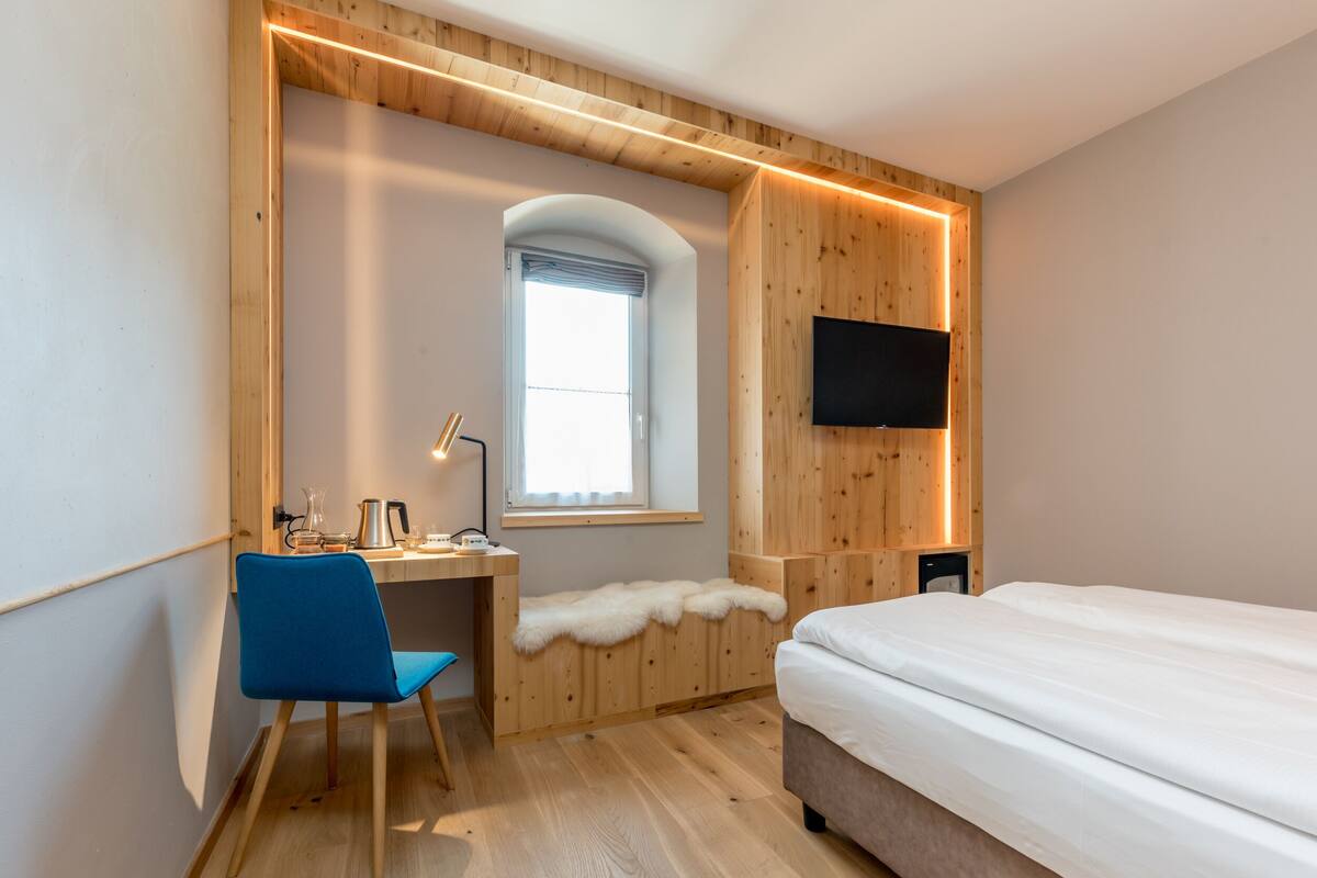A modern hotel room with a wooden accent wall, a desk with a chair, a bed, a television, and a window with a view.