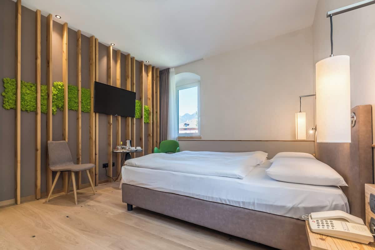 A modern hotel room with a bed, a chair, a TV mounted on a wooden wall, a lamp, and a view of the outdoors.