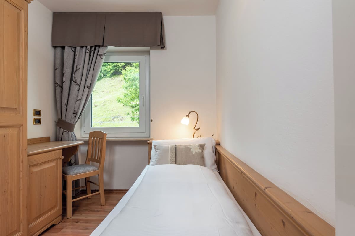 A small bedroom with a single bed, a wooden desk, a chair, and a window with curtains.