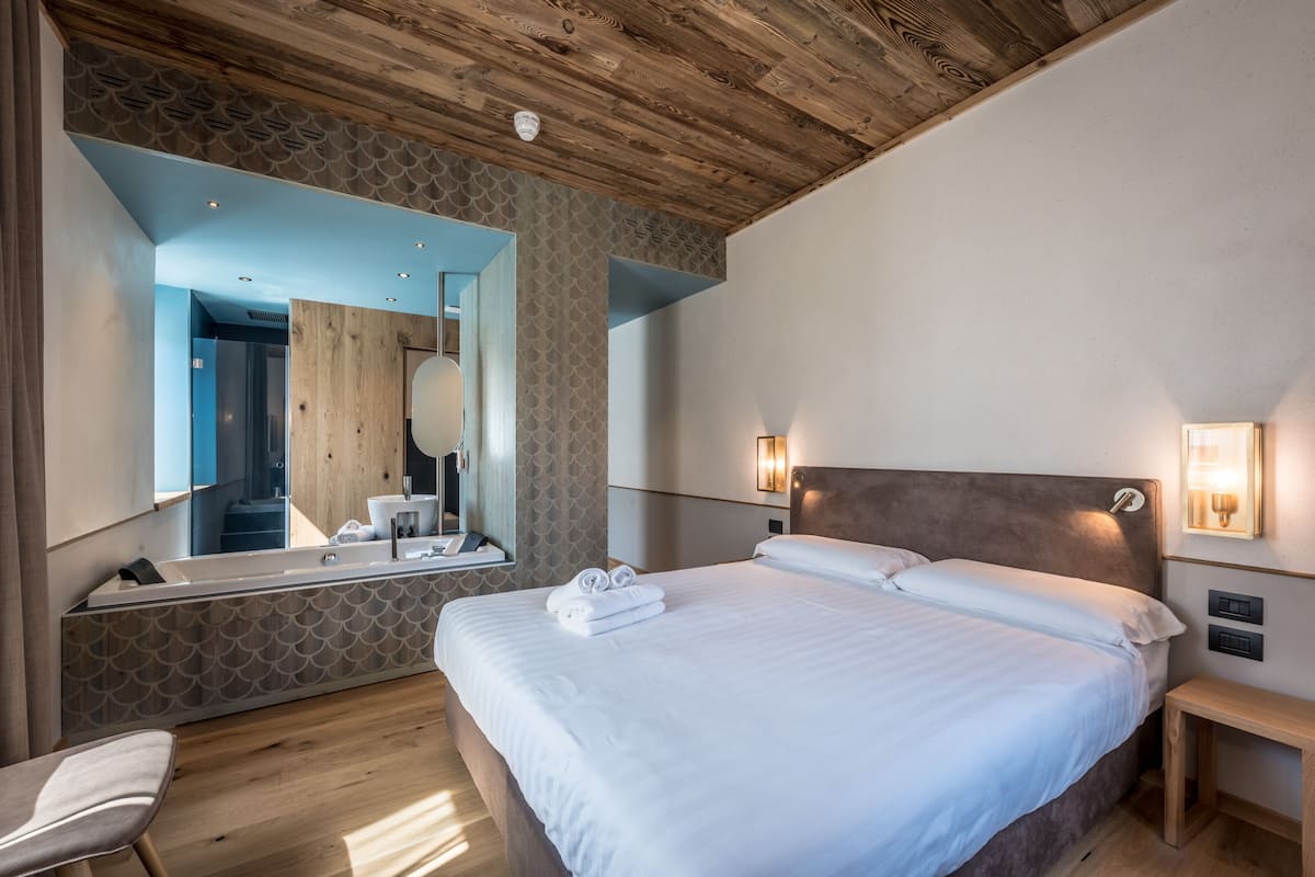 A modern hotel room with a large bed, a wooden ceiling, a large mirror, and a bedside table with a lamp.