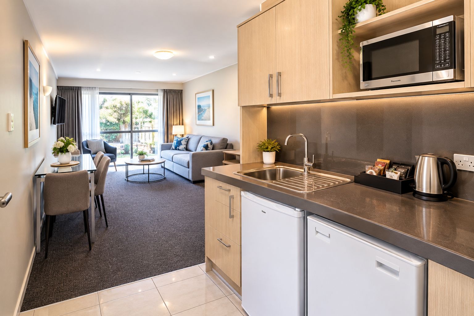 Cocina moderna con microondas integrado, un lavabo y una cafetera. Hay una zona de comedor con una mesa y sillas, y una sala de estar con sofá y una planta.
