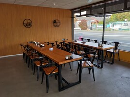A restaurant with long wooden tables and black chairs, large windows with a view of the street.
