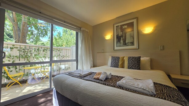 A hotel room with a large bed, a window with a view of greenery, a painting on the wall, and a nightstand with a lamp. - 162 Kings of Riccarton Motel (Christchurch)