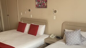 A hotel room with two beds, each with white bedding and red pillows. There is a painting on the wall, a bedside table with a phone, and a red blanket on the floor.