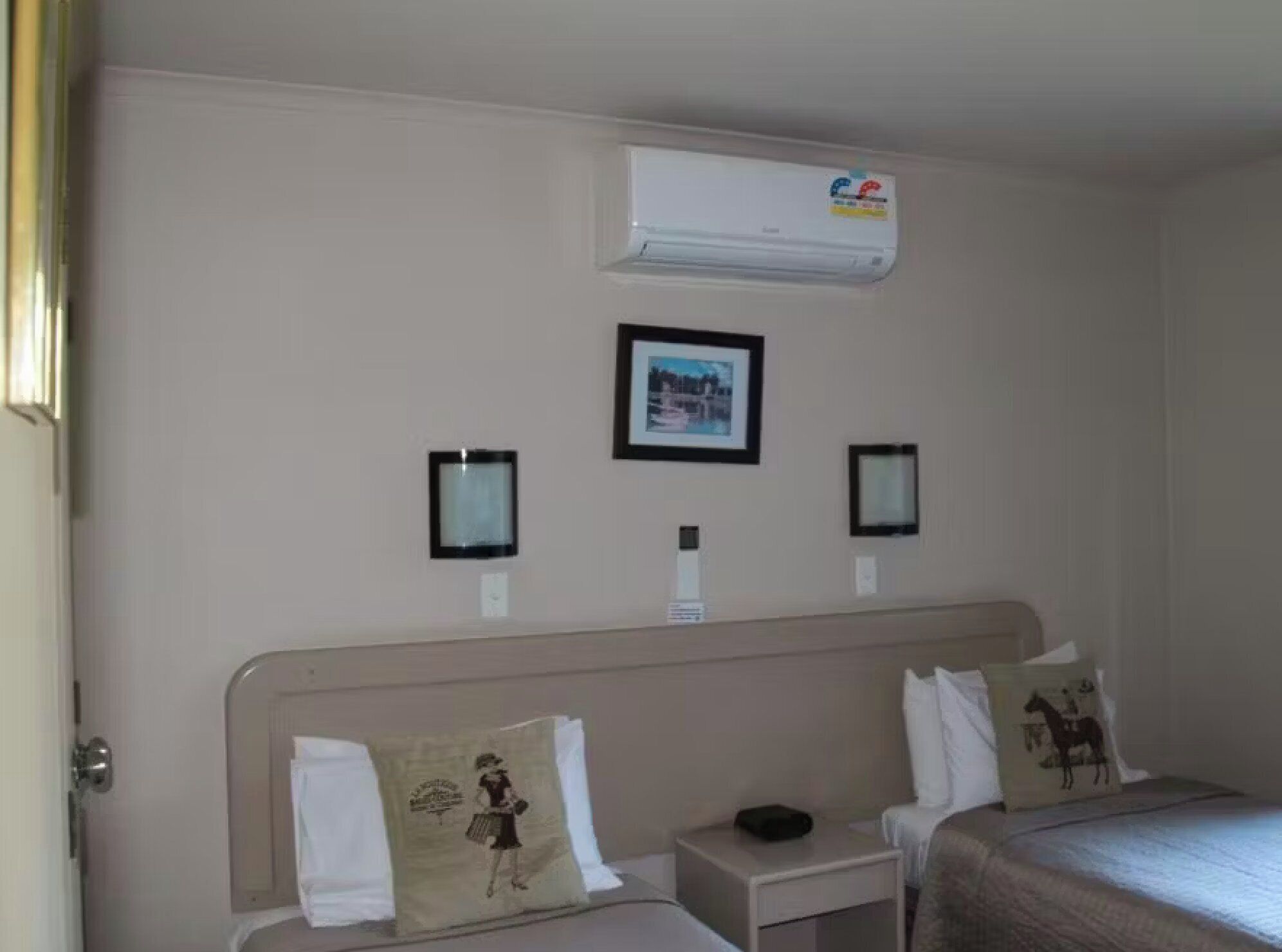 Quarto de hotel com duas camas, uma mesa de cabeceira, ar condicionado fixado na parede e quadros emoldurados.