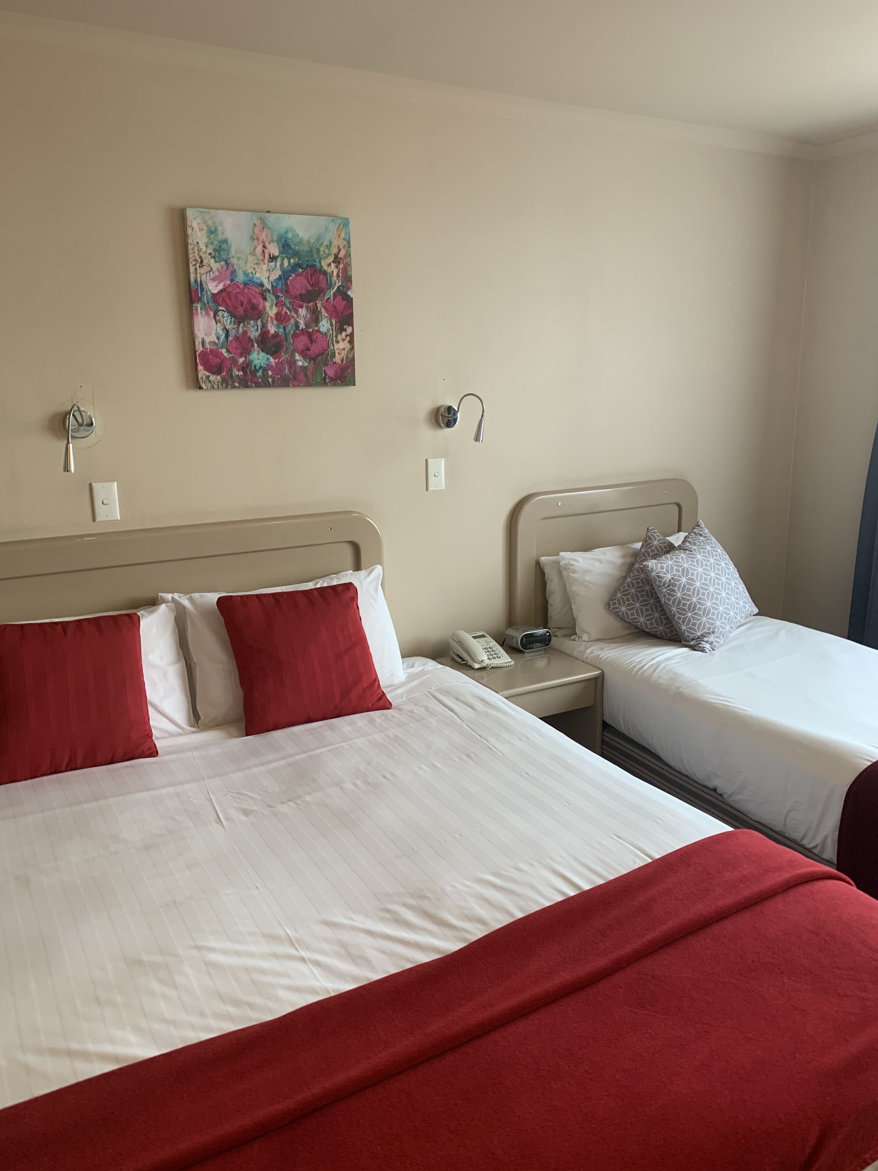 Camera d'albergo con due letti, uno con cuscini rossi e l'altro con cuscini a fantasia. Al muro è appesa una pittura, sul comodino c'è un telefono e tra i letti un tavolino.