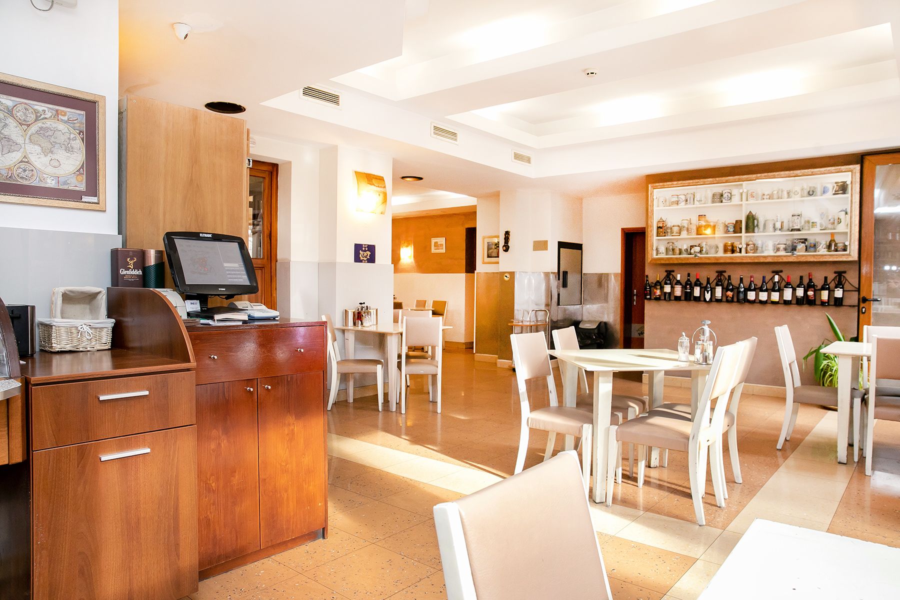 Un hall d’hôtel comprenant un comptoir d’accueil, un bar avec des bouteilles de vin et un espace repas avec des chaises et des tables blanches.