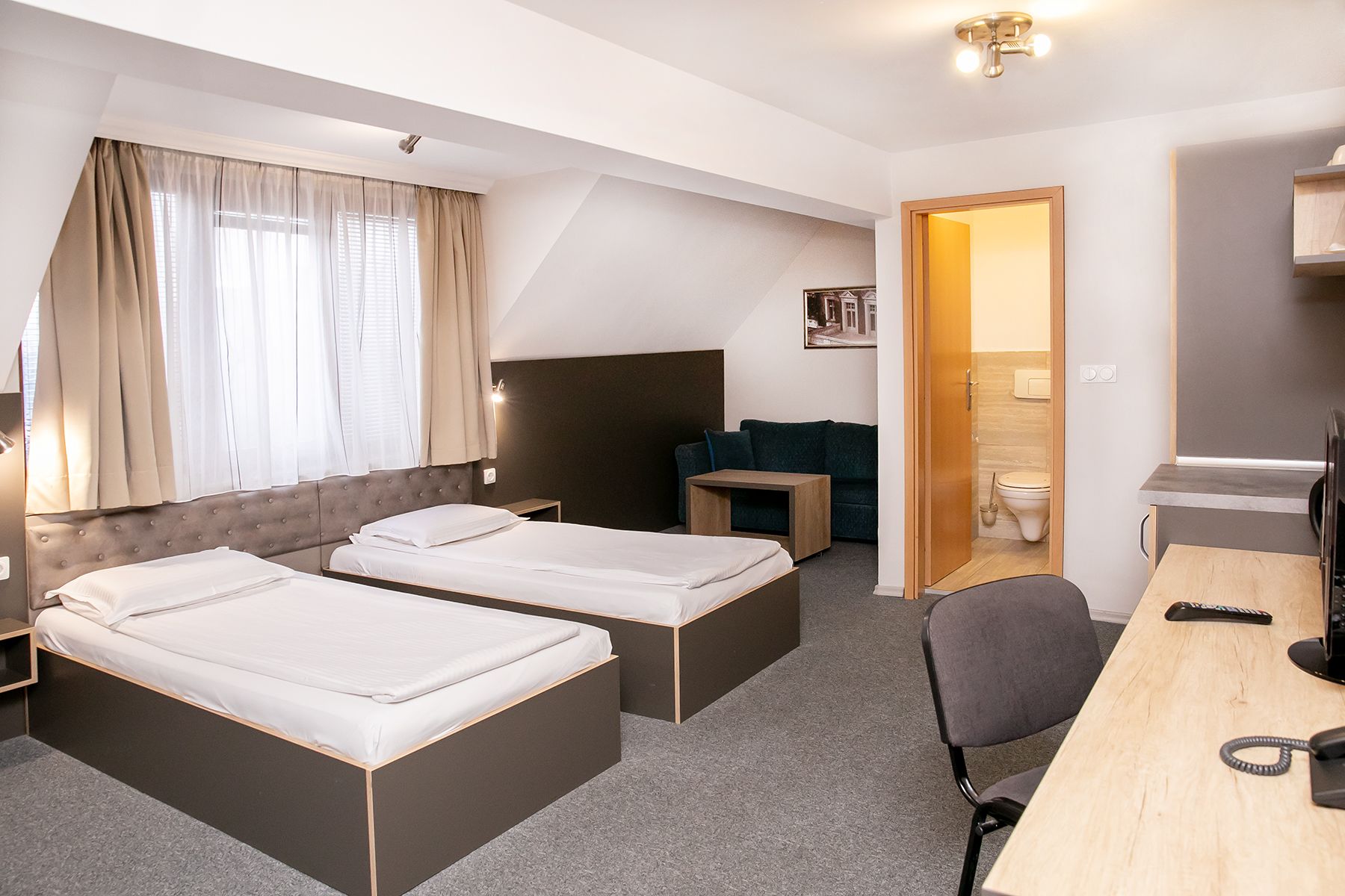 A hotel room with two beds, a desk, a chair, a small table, a sofa, and a bathroom visible through an open door.