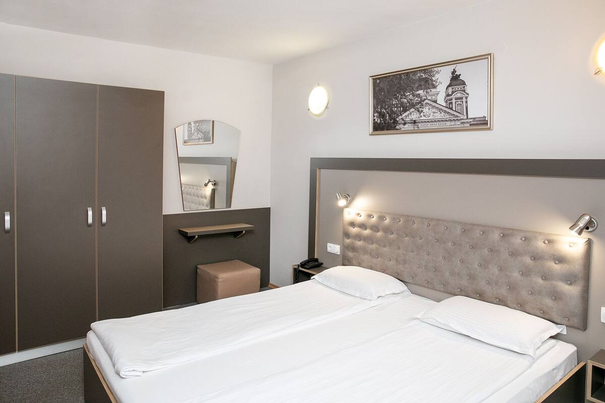 A hotel room with a large bed, a wardrobe, a bedside table with a lamp, a mirror, and a framed picture on the wall.