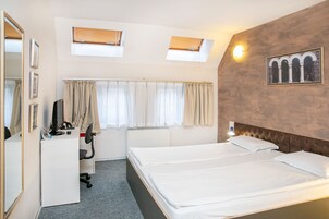 Habitación de hotel con cama, escritorio, silla, espejo, dos ventanas con persianas, una lámpara de pared y un cuadro enmarcado.
