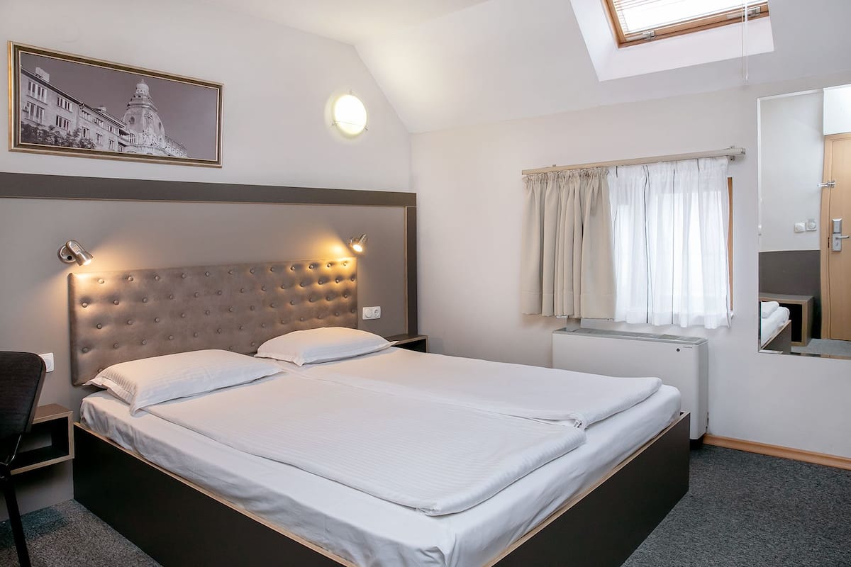 A hotel room with a large bed, a headboard with lights, a window with curtains, a painting on the wall, and a small desk with a chair.