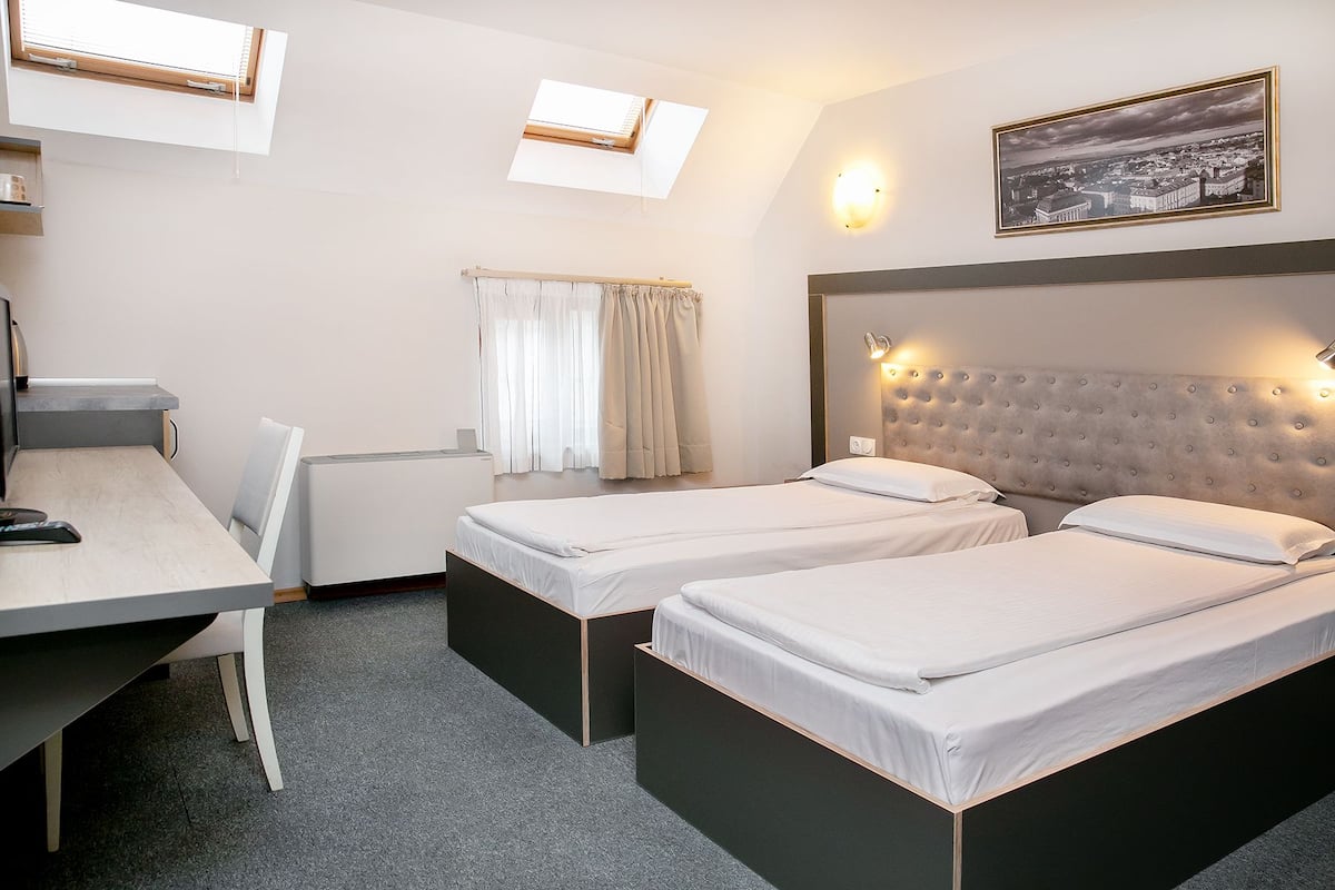 A hotel room with two single beds, a desk, a chair, a window with curtains, a wall-mounted light, and a framed picture.