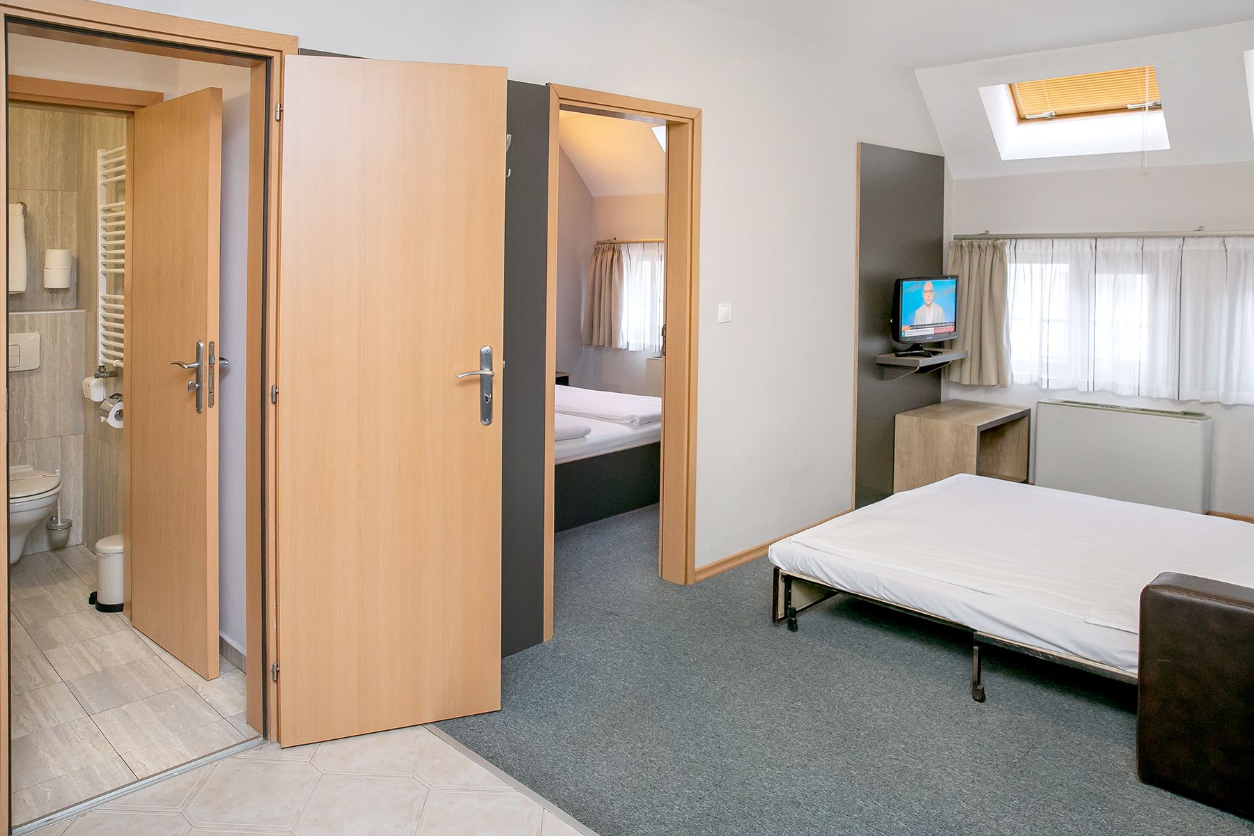 Ein kompaktes Hotelzimmer mit einem Einzelbett, einem kleinen Schreibtisch mit Fernseher, einem Badezimmer mit Toilette und Dusche sowie einem Dachfenster.