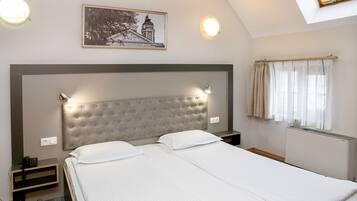 Una habitación de hotel con una cama grande, un cabecero con luces, una mesita de noche pequeña, un teléfono, un cuadro enmarcado en la pared y una ventana con cortinas.