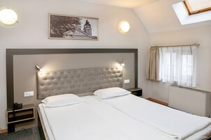 Una habitación de hotel con una cama grande, un cabecero con luces, una mesita de noche pequeña, un teléfono, un cuadro enmarcado en la pared y una ventana con cortinas.