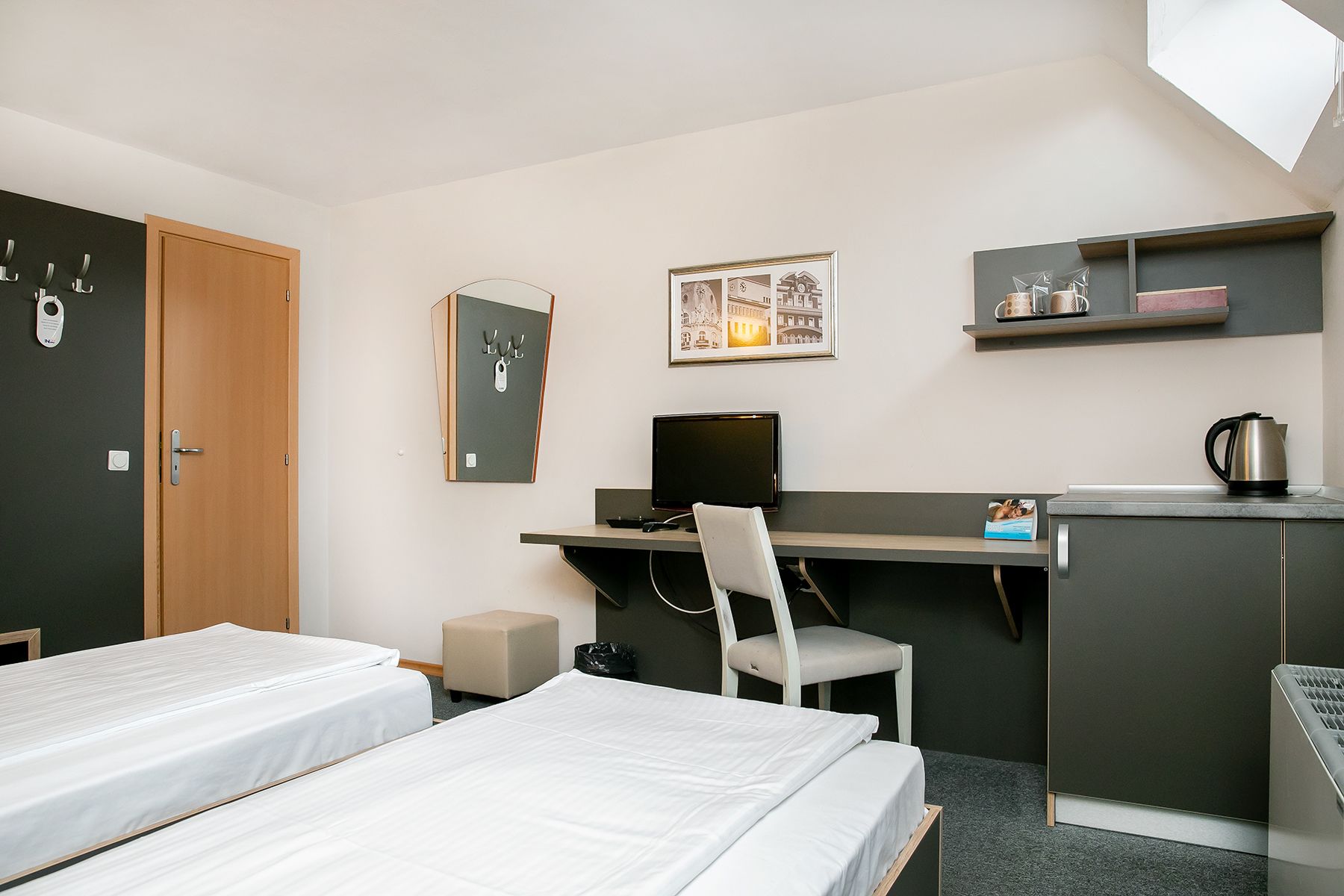 Habitación de hotel con dos camas, un escritorio con una computadora, una silla, un taburete pequeño, un estante con utensilios de cocina y una puerta con un perchero.