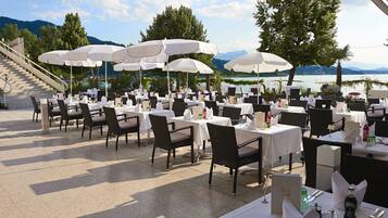 Un espacio de comedor al aire libre con mesas y sillas, sombrillas blancas y vistas panorámicas a un lago.