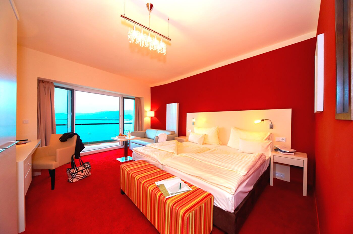 A hotel room with a large bed, a desk, a chair, a small table, and a view of the sea through large windows.
