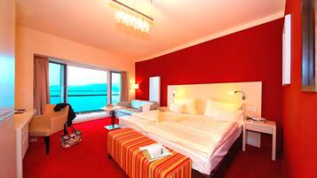 A hotel room with a large bed, a desk, a chair, a small table, and a view of the sea through large windows.