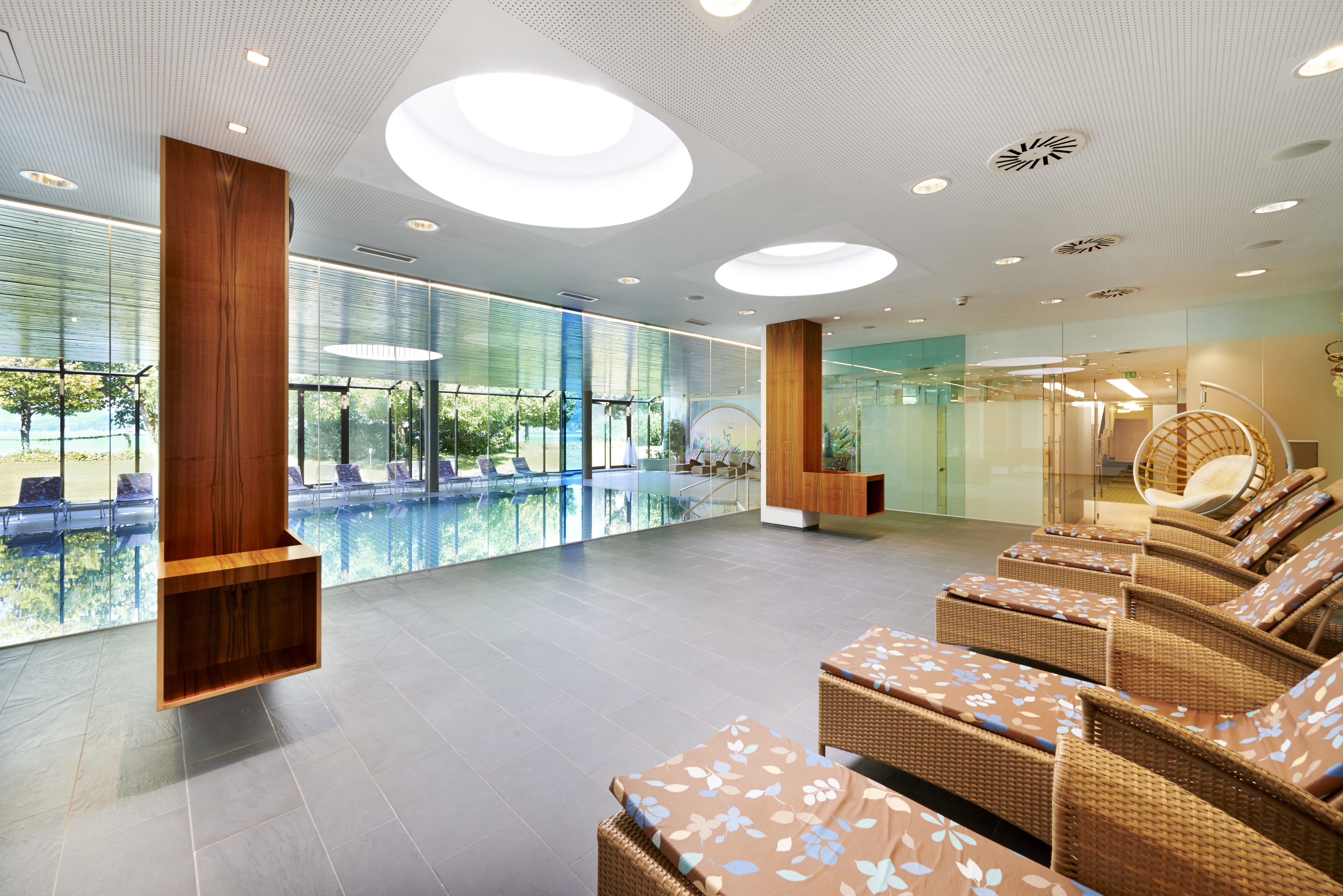 A modern indoor pool area with lounge chairs, wooden columns, and large windows offering views of the pool and outdoors.