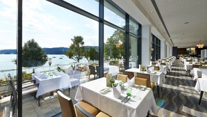 Un restaurant avec des tables dressées pour le service, de grandes fenêtres donnant sur le lac et les collines, et un sol décoré de motifs.