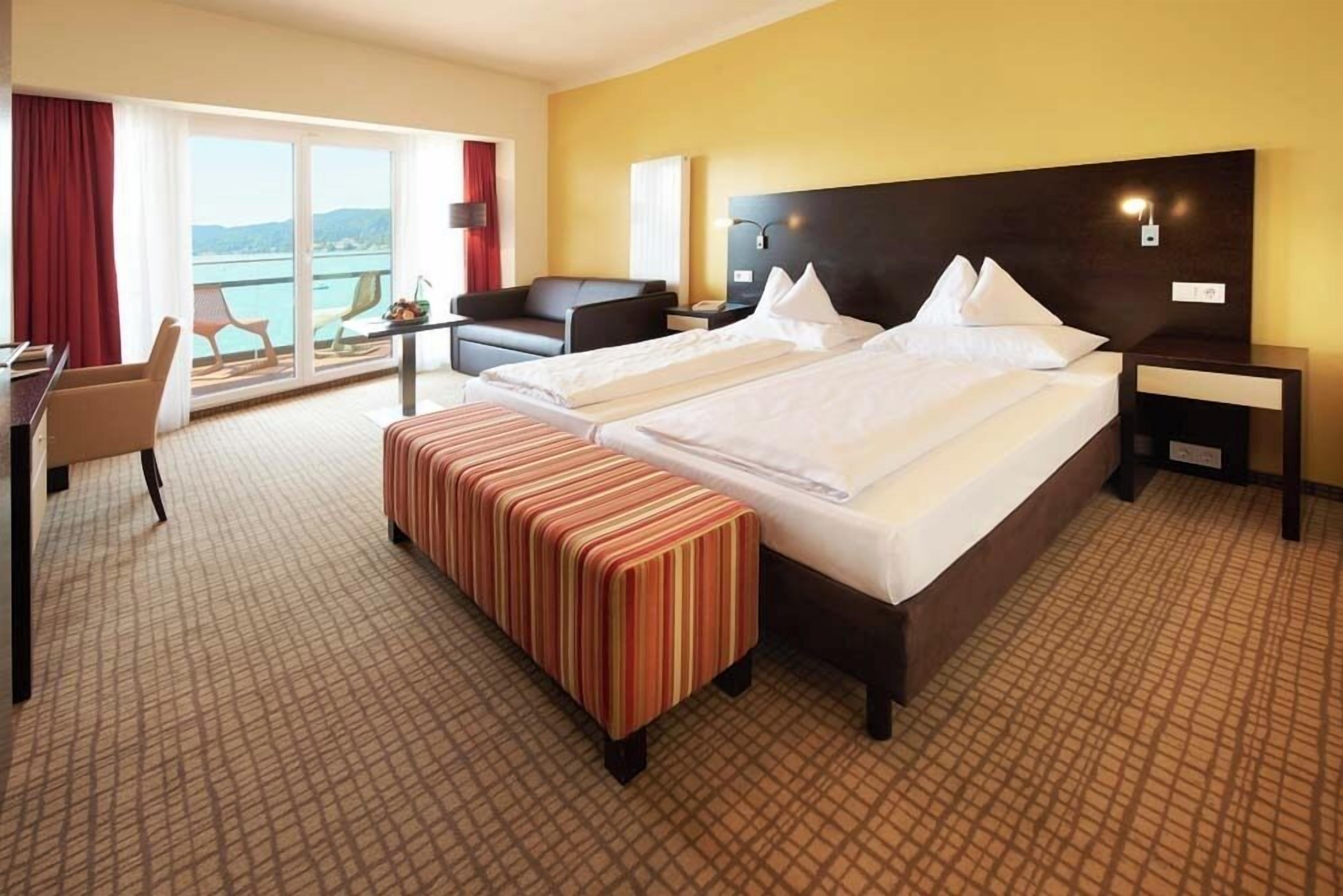 A hotel room with two beds, a desk, a chair, a bench, and a view of the sea.