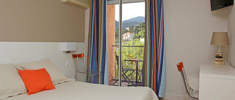 A hotel room with a bed, a desk, a chair, a TV, and a balcony with a view of the mountains.