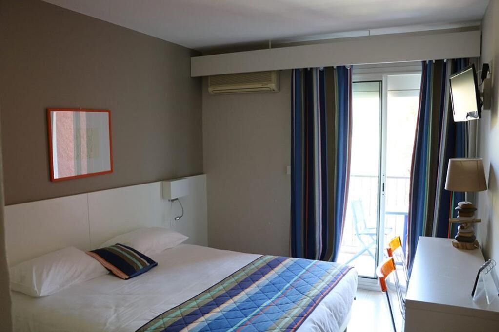 Een hotelkamer met een bed, een nachtkastje met lamp, een raam met gordijnen en een wandairconditioner.